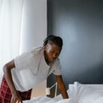 A young black man folding white bed sheets indoors with natural lighting.